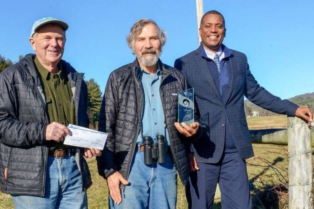 Three men smiling outside with an award of $2,500