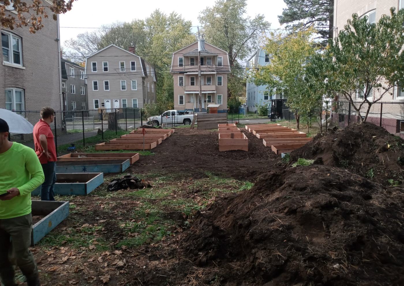 Community garden with 2 gardeners standing on the left, a mound of dirt on the right, and empty raised garden beds in 3 rows