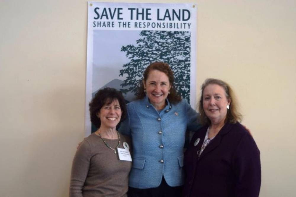 Three women standing in front of a Save the Land poster